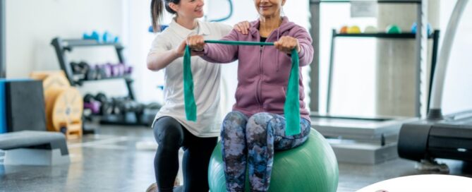 A care provider helps an older woman exercise with a resistance band and an exercise ball, representing how exercise can help with senior fall prevention.