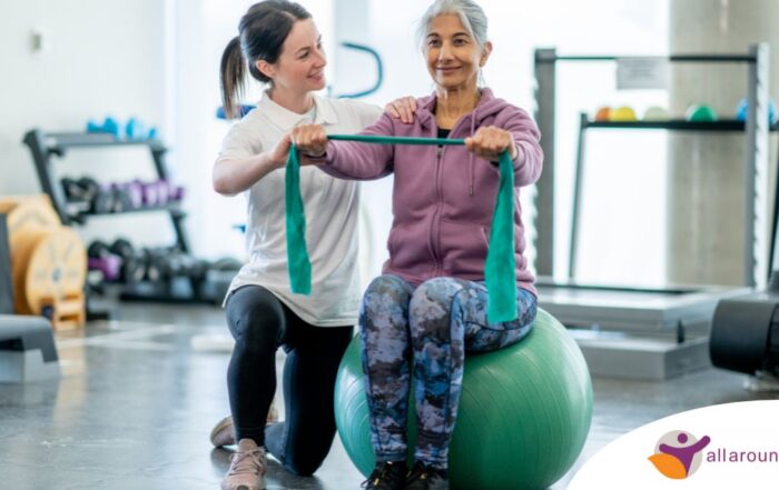 A care provider helps an older woman exercise with a resistance band and an exercise ball, representing how exercise can help with senior fall prevention.